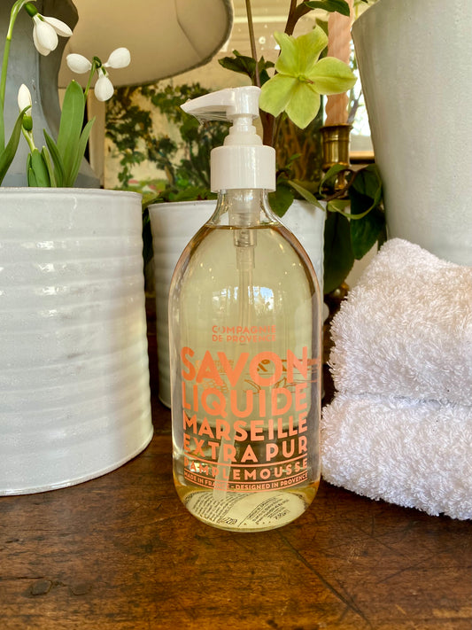 Bottle of Savon Liquide Marseille Extra Pur on a wooden surface with plants and a towel in the background.