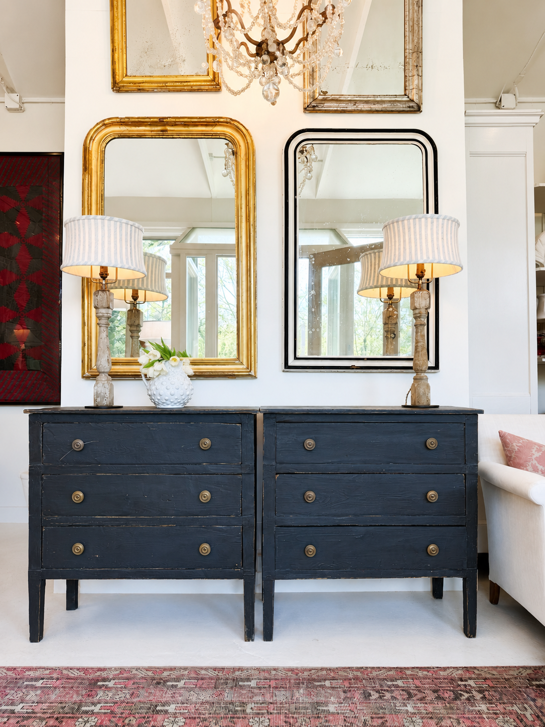 Pair of dark blue dressers with gold knobs in a room with mirrors and lamps.