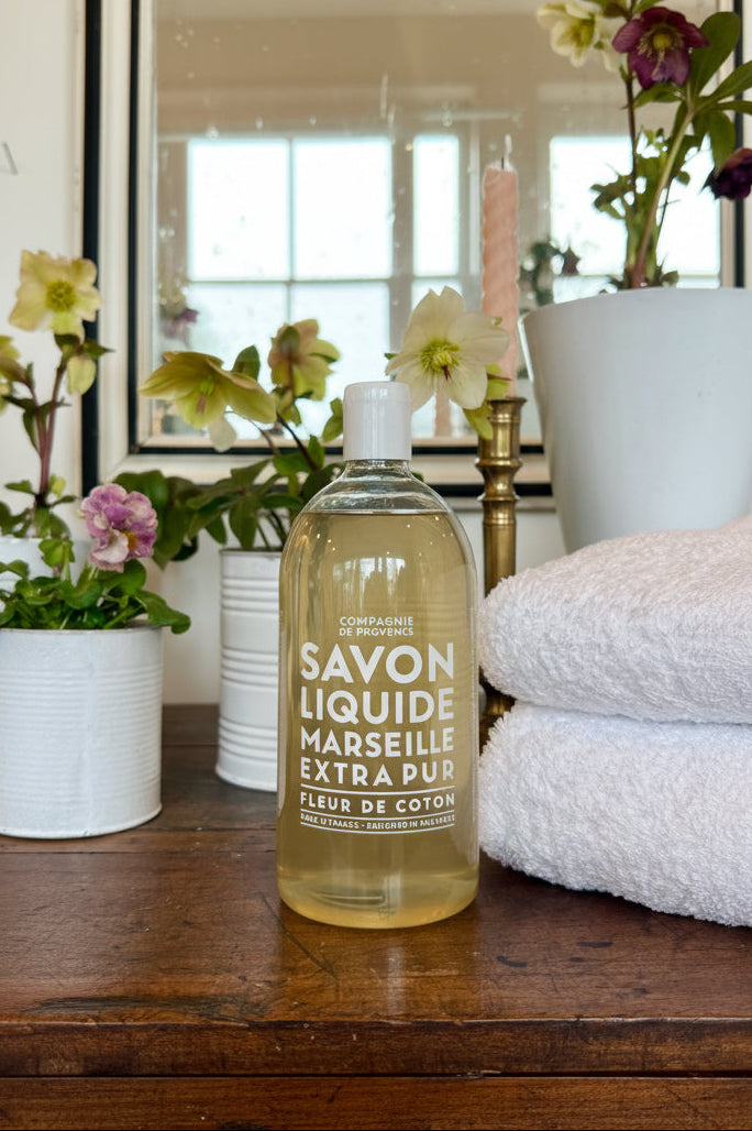 Bottle of Marseille Liquid Soap on a wooden surface with flowers and towels in the background