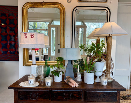 Decorative table with lamps, candles, and plants in a room with mirrors and artwork.