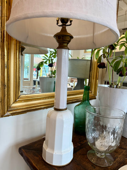 White lamp with a gold base on a wooden surface, with a mirror and decorative items in the background.