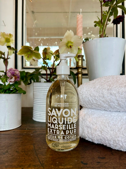 Bottle of Savon Liquide Marseille Extra Pur with flowers and towels in the background