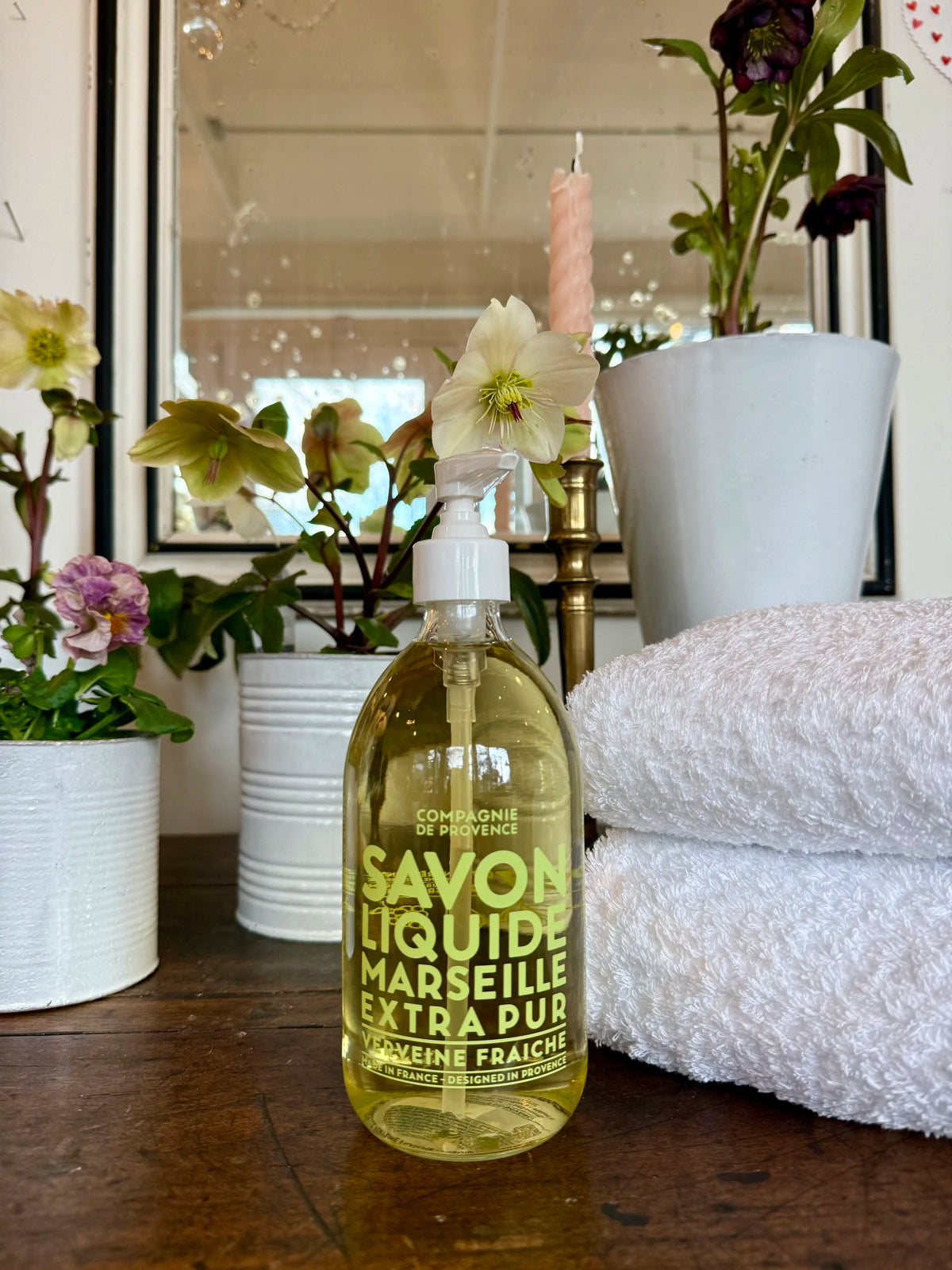 Bottle of Savon Liquide Marseille Extra Pur on a wooden surface with flowers and a mirror in the background.