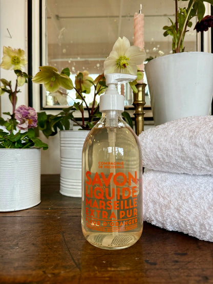 Bottle of Savon Liquide Marseille Extra Pur on a wooden surface with flowers and towels in the background.