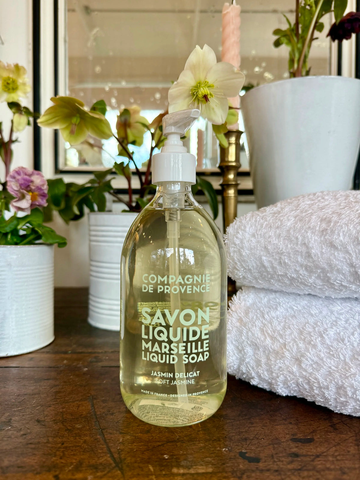 Bottle of Compagnie de Provence liquid soap on a wooden surface with flowers and towels in the background.