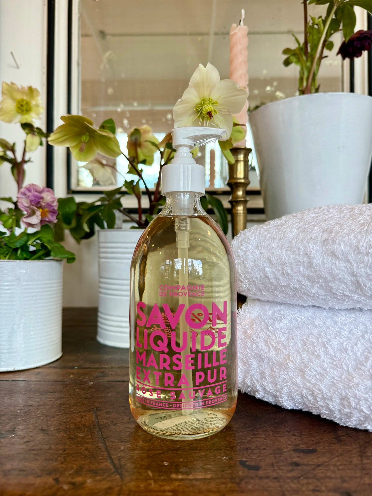 Bottle of Savon Liquide Marseille Extra Pur on a wooden surface with flowers and towels in the background.