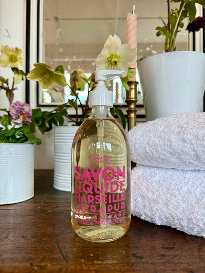 Bottle of Savon Liquide Marseille Extra Pur on a wooden surface with flowers and towels in the background.