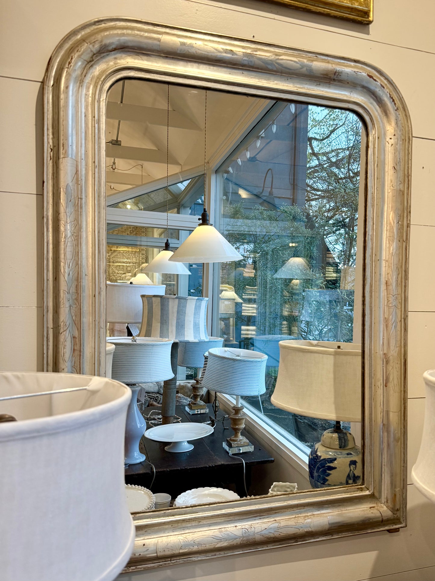 Decorative mirror with a silver frame reflecting a room with lamps and furniture.