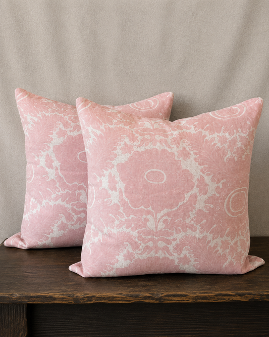 Two pink patterned pillows on a wooden surface with a beige curtain background