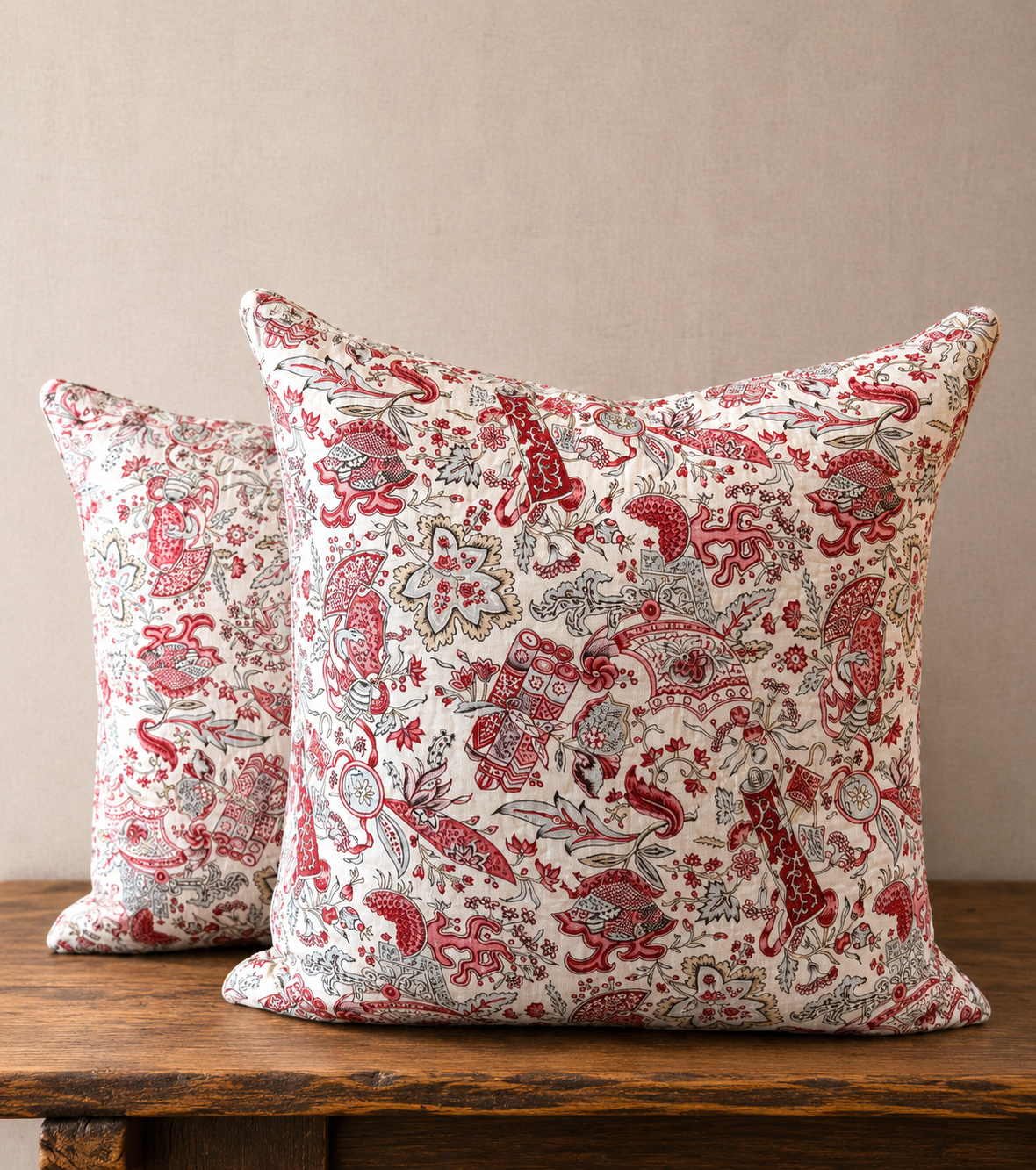 Two decorative pillows with red and beige pattern on a wooden surface against a beige wall.